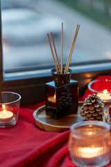 holidays, decoration and celebration concept - aroma reed diffuser and candles on red tablecloth on window sill at home