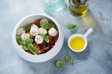 Bowl of caprese salad made with mini mozzarella balls, red cherry tomatoes and fresh green basil, elevated view on a light-blue stone surface