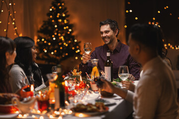 holidays, party and celebration concept - multiethnic group of happy friends having christmas dinner at home and drinking wine