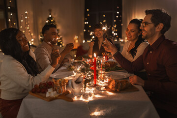 holidays and celebration concept - multiethnic group of happy friends with sparklers having christmas dinner at home