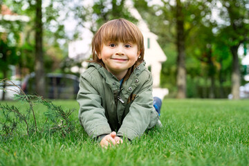Portrait of a little preschool boy wearing green parka lying on the lawn in fresh day. Happy smiling child outdoor.
