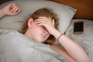 A teenage girl is sleeping in her bed, covering her face with her hand. There is a phone next to the pillow. Close-up. Awakening, day mode, morning concept