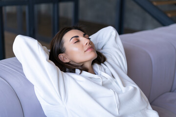 Young woman at home in a white hoodie on the couch serenely sits with closed eyes, relaxes, rests, meditates