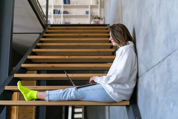 A young woman at home in a white hoodie and jeans sits on the stairs with a laptop on her knees, works remotely, studies