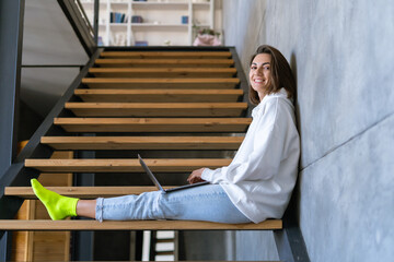 A young woman at home in a white hoodie and jeans sits on the stairs with a laptop on her knees, works remotely, studies