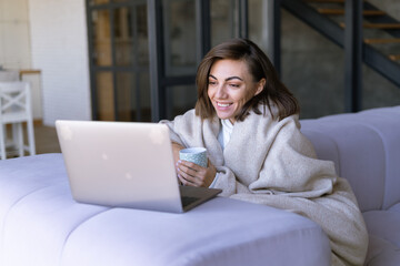 Young woman at home on a sofa in winter under a cozy blanket with a laptop, watching comedy series, laughing, smiling in a winter evening