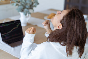Obraz premium Young woman at home in the kitchen in a white hoodie with a laptop, a graph of growth on the screen, rejoices the cryptocurrency is growing up