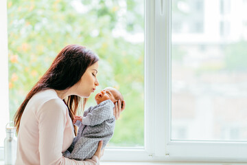 Mother Gently Kissing Cheek of Newborn Baby in Hospital. Portrait of mom hugging and kissing her newborn baby son against big window. Loving and caring parents with little children concept.