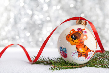 Christmas decoration - a hand-painted ball with the symbol of the Year of the Tiger. Nearby is a New Year tree branch and a red ribbon. Blurred background of silver color