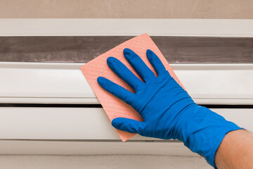 The hand of a man in a blue rubber household glove wipes and cleans the air conditioner. Maintenance and cleaning indoor service