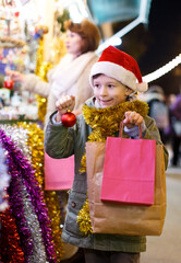 Fototapeta na wymiar Portrait of happy tween boy holding paper bags with Christmas decorations with mother behind on outdoors fair