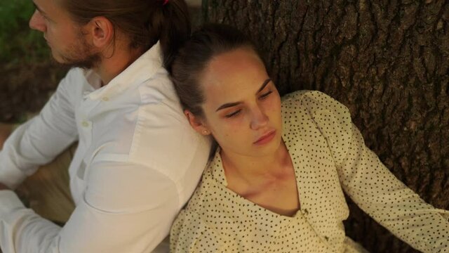 Man white shirt, beige shorts sits back girl long hair beige sundress. Husband wife, carefree, peaceful, sit under a tree trunk shade national park their backs each other, enjoying moment nature air