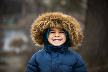 Portrait of preschool smiling boy in warm coat with hood in winter outdoors