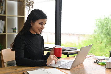 Photo of young creative woman holding a red coffee cup while using a computer laptop at the wooden...