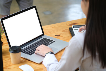 Behind view of a young accountant working with a white screen computer laptop and calculator at the wooden table surrounded by a coffee cup and various equipment.