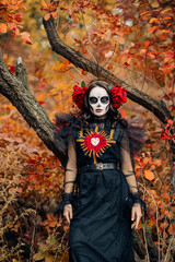 Woman with sugar skull makeup dressed as Santa Muerte is against background of autumn forest.