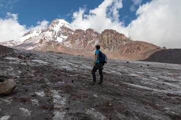 Fototapeta premium A man standing at the foothill of Gergeti Glacier, with the view on Mount Kazbeg, Caucasus, Georgia. Barren slopes below the snow-capped peak. Few clouds gathering around the sharp peak. Tranquillity