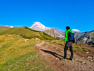 A man enjoying the view on a cloudless view on Mount Kazbeg in Caucasus, Georgia. There are lush pastures and green hills below the snow-capped peak of the glacier. Serenity and Tranquillity. Nature
