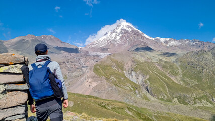 A man leaning agains a stony pillar and enjoying the view on a cloudless view on Mount Kazbeg in Caucasus, Georgia. There are lush pastures and green hills below the snow-capped peak of the glacier. © Chris