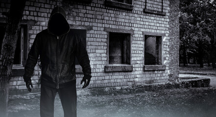 Photo of scary horror stranger stalker man in black hood and clothing on abandoned building background.