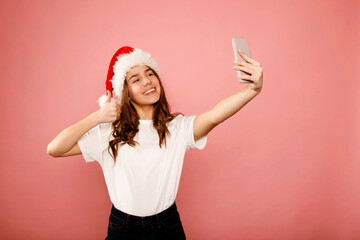 Happy caucasian girl teenager standing in white t-shirt and santa claus red hat holding smartphone doing selfie with hand ok gesture over pink background. Studio portrait. Christmas and New year Holid