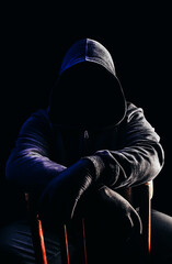 Photo of scary horror stranger stalker man in black hood and clothing sitting on chair on dark...