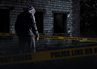 Photo of scary horror stranger stalker man in black hood and clothing on abandoned building background with police line stripes.