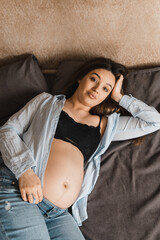 Young brunette pregnant woman having rest in the bed. Sexy pregnant female in the bedroom. Pregnant woman in blue jeans and husband's shirt.Selective focus. Film grain.