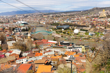 Fototapeta premium Scenic view of Tbilisi Old town on banks of Kura river on sunny spring day overlooking modern pedestrian Bridge of Peace, Music Theater building and air balloon in Rike Park, Georgia