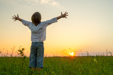 Happy boy with raised hands looks at the sunset  in summer. Boy in a field with his hands raised...