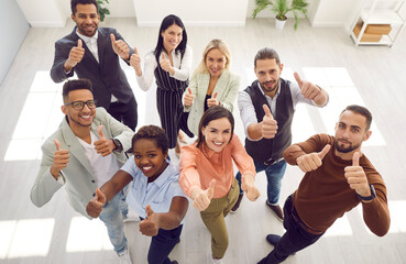 Fototapeta na wymiar Diverse team of happy motivated young mixed race business people standing in office, looking up, smiling and giving thumbs up all together. High angle, from above, top view. Teamwork, success concept