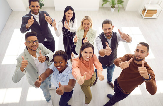 Diverse team of happy motivated young mixed race business people standing in office, looking up, smiling and giving thumbs up all together. High angle, from above, top view. Teamwork, success concept