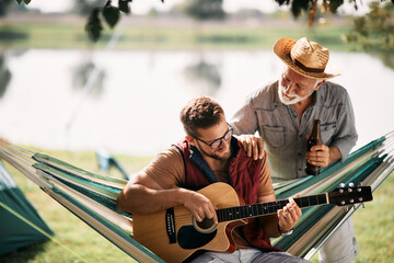 Smiling man plays acoustic guitar while camping with his senior father by lake.