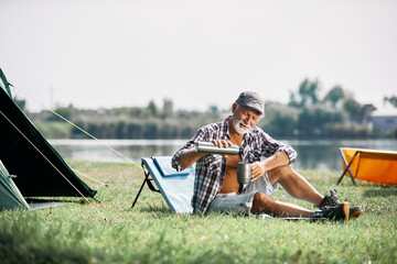 Senior man uses water from thermos while relaxing on grass and camping.