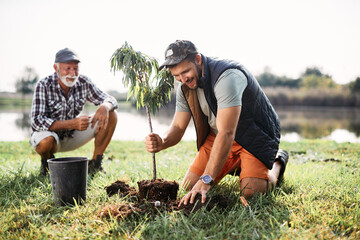 Happy man plants tree sapling with his father by the lake.