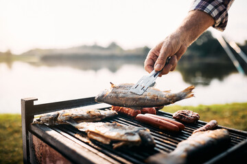 Close-up of man grills fish on barbecue grill while camping by river.