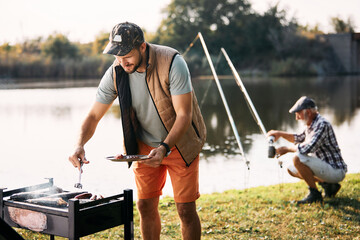 Happy man grills fish and meat on barbecue grill after fishing with his father in nature.