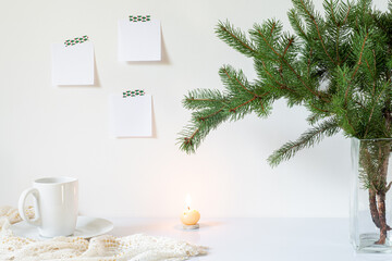 Festive breakfast still life.Cup of coffee, candle and vase with fir branch.Empty notepads mockup hanging on the white wall.Christmas decor.Working space, home office.Happy new year resolutions, list
