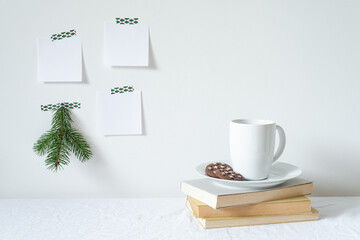 Festive breakfast still life.Cup of coffee with cookies, books on desk.Fir branch, empty notepads mockup hanging on the wall.Christmas decor.Working space, home office.Happy new year resolutions, list