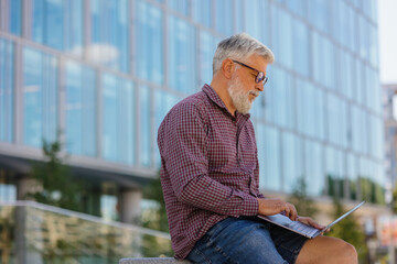 an adult gray-haired man with a beard is sitting at the office and working on a laptop. a senior businessman in a shirt and with a laptop is making a career in a large company. a stylish and