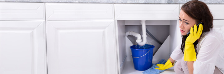 Woman Calling Plumber In Front Of Water Leaking From Pipe