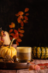 Pumpkin spicy latte in a stylish glass on a dark background with pumpkins and autumn leaves. Autumn composition in warm colors