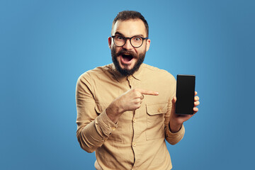 Portrait of surprised young man wearing beige shirt and eyeglasses, isolated over blue background, showing blank screen mobile phone 