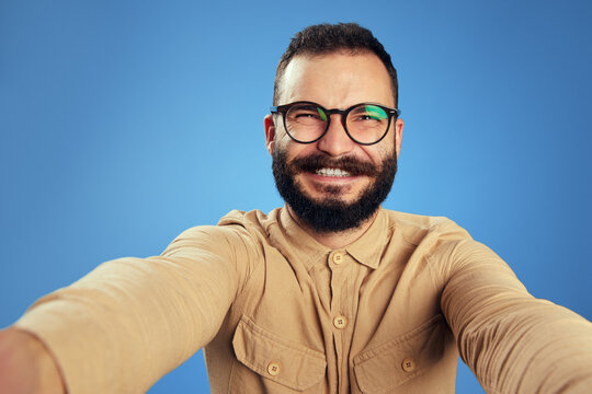 Cheerful bright bearded man in beige shirt and eyeglasses laughing at camera taking selfie on blue background