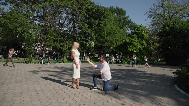 man gets down on one knee and makes an offer with ring in box in his hands to beautiful young woman, background of crowd of people running towards couple in love, shouting no