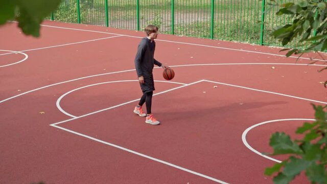 Side view wide slow motion shot of teenage boy training basketball handling and dribbling on outdoor college court. Young man, college or university student, practicing basketball alone