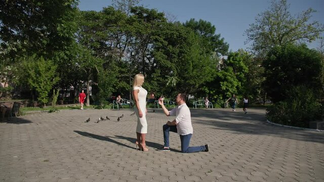 marry me, loving man on his knees proposes with help of wedding ring to handsome young woman background of couple of people running towards park in park