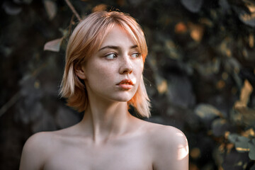 a young blonde without clothes looks towards a portrait in a park against a background of foliage