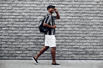 African American man in a summer shirt and shorts, steps against a black brick wall, black guy