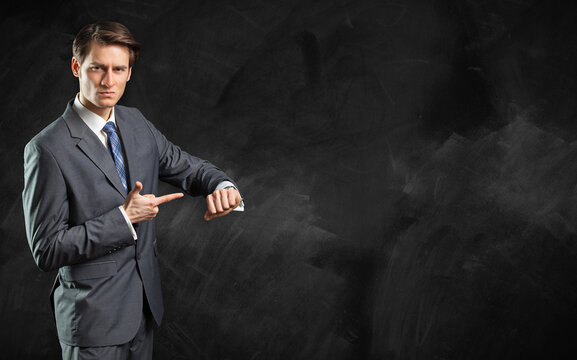 businessman standing in front of a blackboard pointing at his watch as a concept for an upcoming deadline